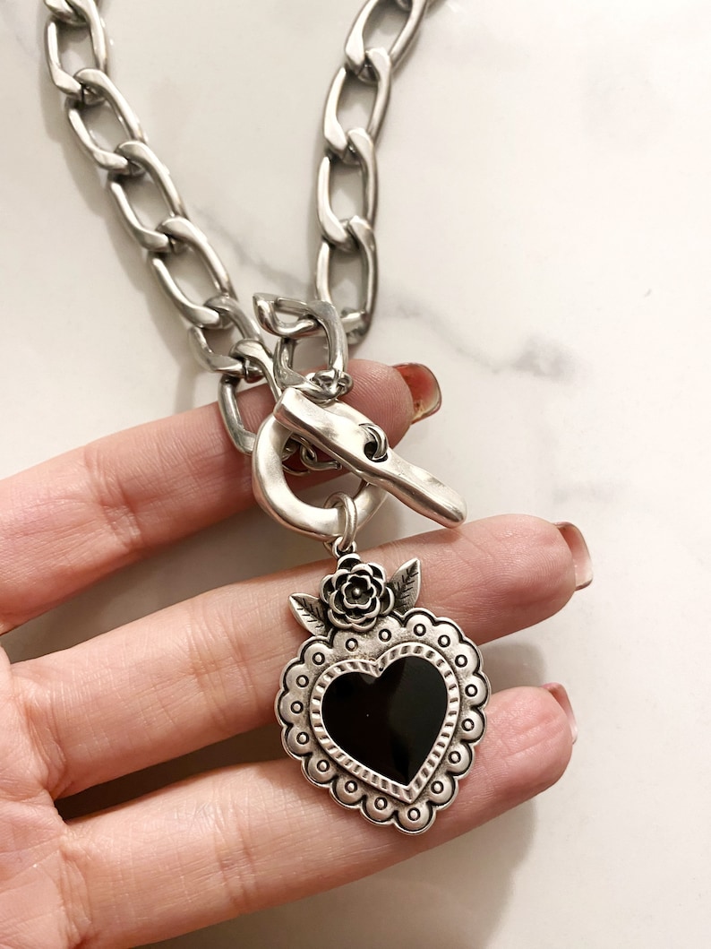 May include: Silver chain necklace with a large silver heart pendant. The heart has a black center and is decorated with a silver flower and leaves.