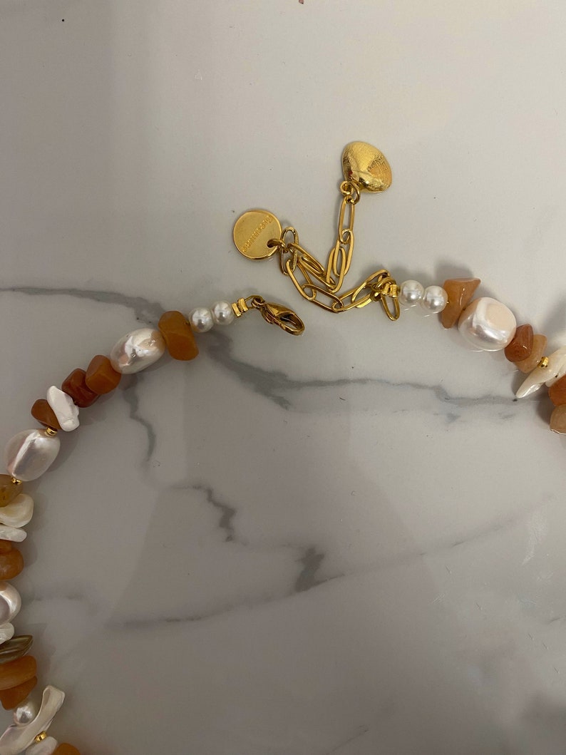 May include: A gold chain necklace with a brown and white stone bead design. The necklace has a small gold charm with the word "Handmade" on it.