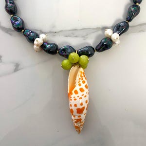 May include: A black pearl necklace with a seashell pendant. The pendant is a white and orange seashell with brown spots. The necklace is accented with small white pearls and green beads.