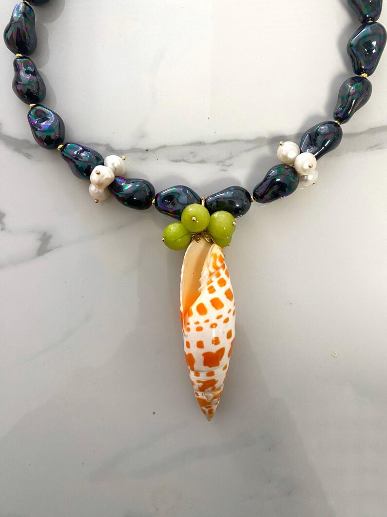 May include: A black pearl necklace with a seashell pendant. The pendant is a white and orange seashell with brown spots. The necklace is accented with small white pearls and green beads.
