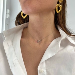 May include: A woman wearing a white button-down shirt with a delicate gold necklace and large gold heart-shaped earrings.
