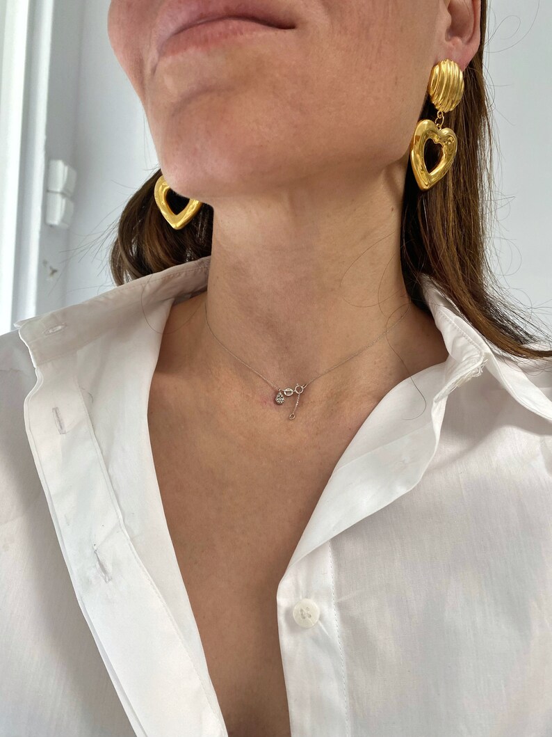 May include: A woman wearing a white button-down shirt with a delicate gold necklace and large gold heart-shaped earrings.