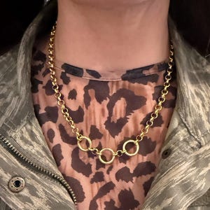 Peut inclure: Un collier à chaîne doré composé d'une série de maillons circulaires uniformes reliés par un maillon circulaire central plus grand. Le collier est porté sur un haut à motif léopard marron et noir et une veste camouflage grise à bouton.