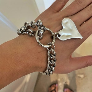 Könnte beinhalten: Ein silbernes Kettenarmband mit einem großen Herz-Anhänger. Das Armband besteht aus zwei verschiedenen Kettenarten, einer mit kleinen runden Gliedern und einer mit größeren, massiveren Gliedern. Der Herz-Anhänger ist mit einem großen, runden Ring am Armband befestigt.