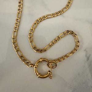 May include: A gold-toned figaro chain necklace featuring a circular spring ring clasp. The chain consists of a repeating pattern of three small circular links followed by one elongated oval link, connected to a round, polished metal fastener.