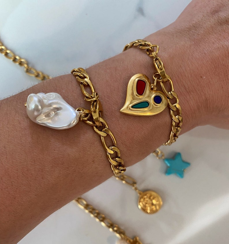 May include: Gold chain bracelet with multiple charms, including a heart with red, blue, and green enamel, a large white pearl, a blue star, and a gold coin.