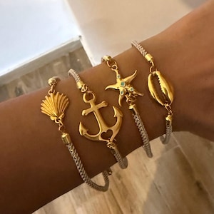 May include: Four gold-toned nautical charm bracelets. Each bracelet features a different charm: a seashell, an anchor, a starfish, and a seashell. The bracelets have a woven, silver-colored band.