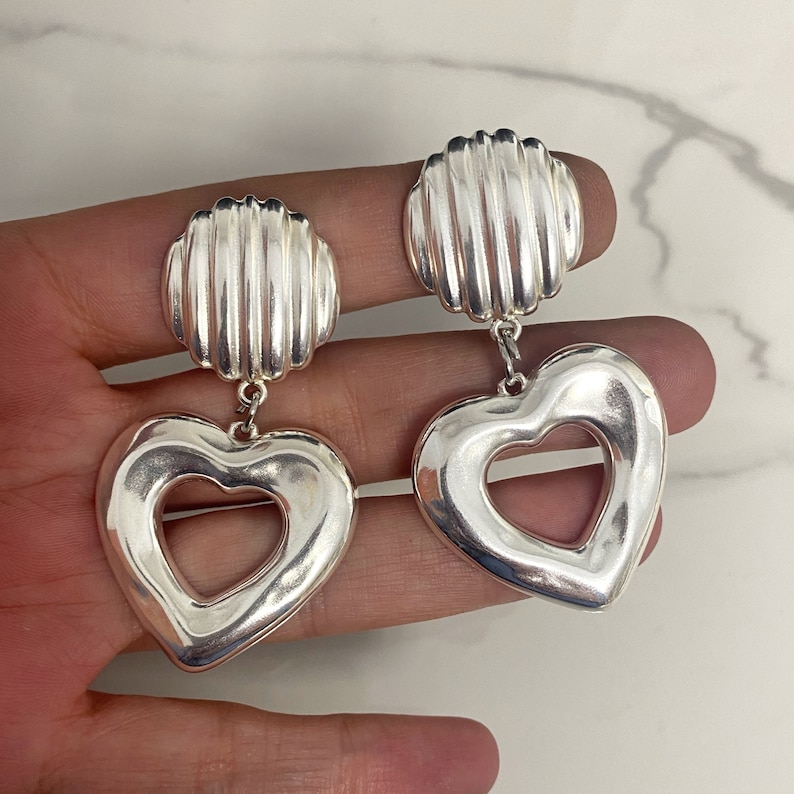 May include: Silver heart shaped earrings with a textured round top. The heart has a cutout in the center.