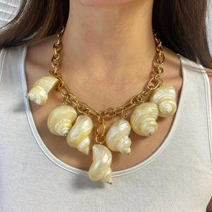 Op de afbeelding: Een gouden ketting met meerdere grote witte schelpen eraan bevestigd. De schelpen zijn iriserend en hebben een natuurlijke, organische textuur.