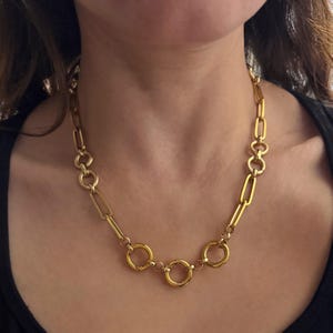 May include: A gold-colored necklace with a variety of link styles. The necklace features a mix of elongated and circular links, creating a bold and stylish accessory. The necklace is worn around the neck.