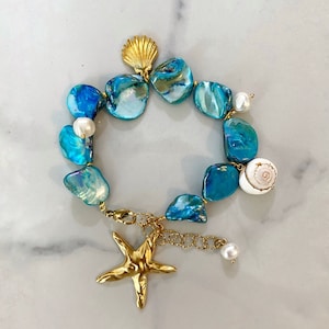 Könnte beinhalten: Ein blaues und goldenes Muschel-Armband mit einem Seestern-Anhänger. Das Armband besteht aus schillernden blauen und grünen Muschelteilen und hat einen goldenen Seestern-Anhänger, der daran hängt. Das Armband ist perfekt für einen sommerlichen Look.