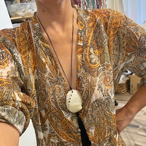 May include: Abalone shell pendant necklace with a black tassel. The necklace features a large, iridescent abalone shell suspended from a black cord. The shell is cream-colored with natural markings. The background is a paisley print blouse in shades of orange, brown, and cream.