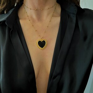 Pu&ograve; includere: Una collana a catena dorata con un ciondolo a forma di cuore nero. Il ciondolo a forma di cuore ha un design dettagliato con un bordo smerlato.