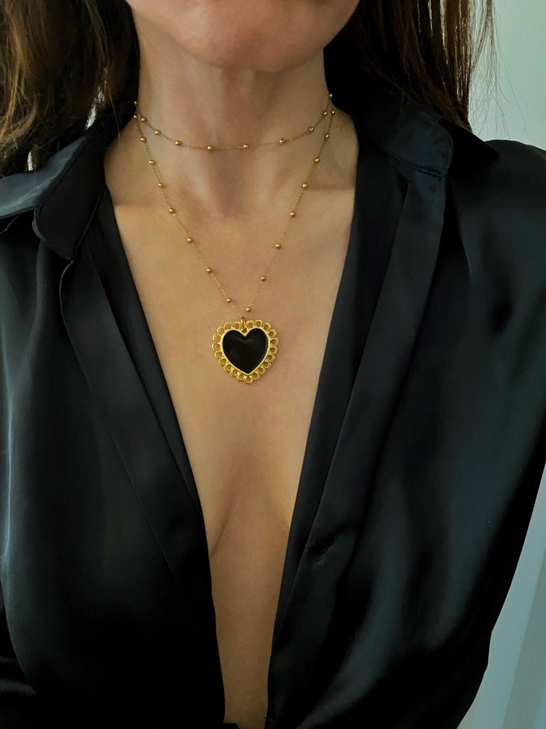 Pu&ograve; includere: Una collana a catena dorata con un ciondolo a forma di cuore nero. Il ciondolo a forma di cuore ha un design dettagliato con un bordo smerlato.