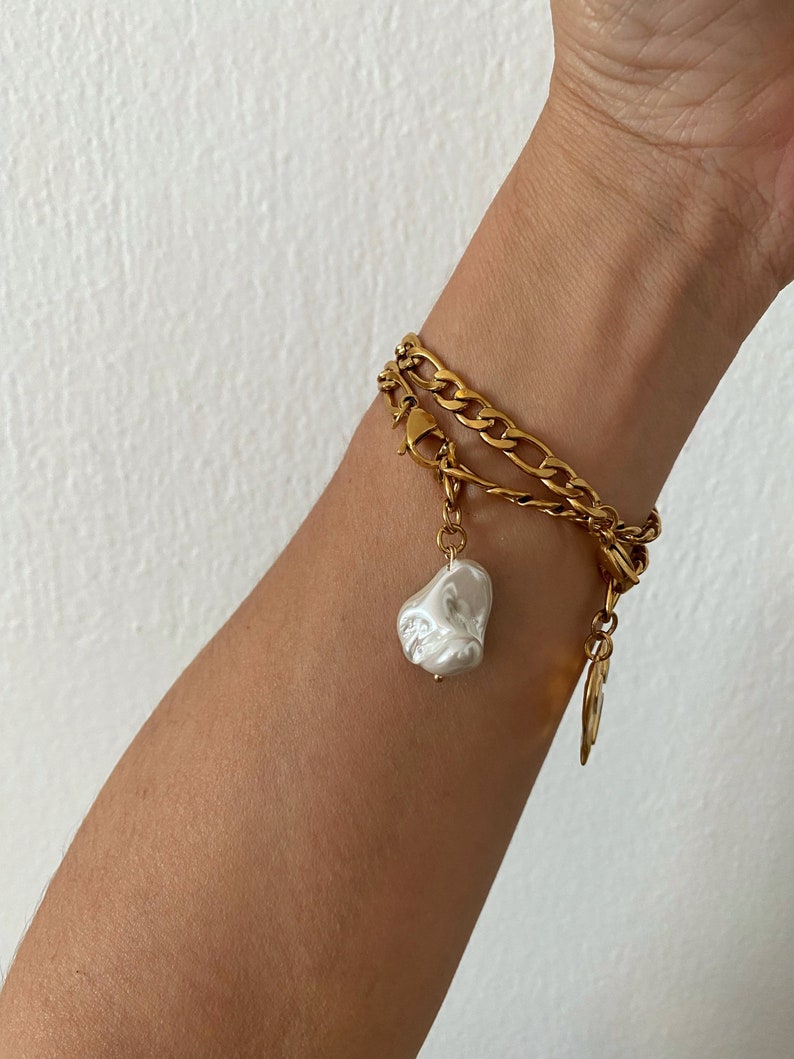 May include: A gold chain bracelet with a large white pearl charm. The bracelet is layered with two different chain styles.