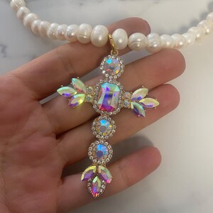 Può includere: Una collana d'oro con un grande ciondolo a forma di croce in cristallo iridescente e una fila di perle bianche.