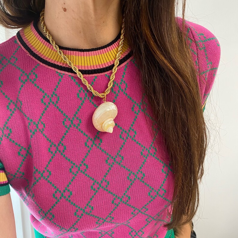 May include: A gold chain necklace with a large seashell pendant. The necklace is worn by a person wearing a pink and green patterned sweater.