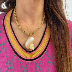 May include: A gold rope chain necklace with a large seashell pendant. The seashell is white with pink and brown stripes. The necklace is worn by a person wearing a pink and green patterned sweater.