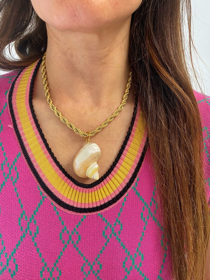 May include: A gold rope chain necklace with a large seashell pendant. The seashell is white with pink and brown stripes. The necklace is worn by a person wearing a pink and green patterned sweater.