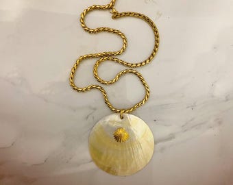 May include: A gold-toned necklace featuring a large, round, iridescent pendant with a small, textured gold seashell accent. The chain is a twisted rope design. The necklace is displayed on a marble surface.