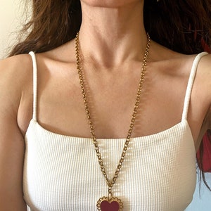 May include: A gold chain necklace with a large red heart pendant. The heart has a scalloped edge and is set in a gold frame. The necklace is worn by a person with long brown hair.