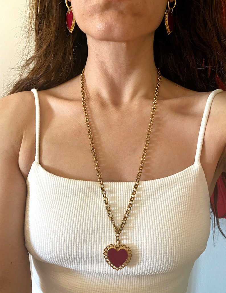 May include: A gold chain necklace with a large red heart pendant. The heart has a scalloped edge and is set in a gold frame. The necklace is worn by a person with long brown hair.
