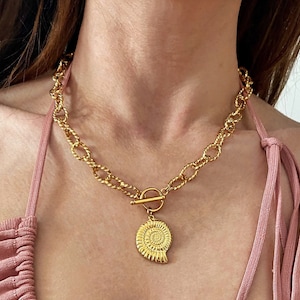 Könnte beinhalten: Eine goldene Kettenkette mit einem großen goldenen Anhänger in Form einer Nautilus-Schale. Die Kette besteht aus dicken, gedrehten Gliedern.
