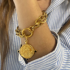Könnte beinhalten: Ein goldenes Kettenarmband mit einem großen, runden, goldenen Anhänger mit filigranen Details. Der Anhänger ist mit einem goldenen Ring an der Kette befestigt.