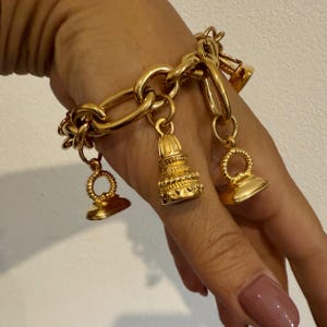 May include: A gold-toned chain link bracelet featuring three dangling charms. The charms include a central bell-shaped piece with intricate ribbed detailing and two smaller, flat, circular-topped seal-style charms hanging from the thick metal links.