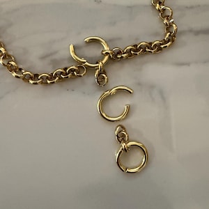 Op de afbeelding: Een goudkleurige ketting met een ronde veerringsluiting. De ketting bevat twee extra losse ronde veerringonderdelen, waarvan er één een kleine springring aan de basis heeft, allemaal gemaakt van gepolijst metaal.