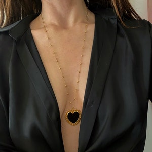 Pu&ograve; includere: Una collana con catena dorata con un ciondolo a forma di cuore nero. Il ciondolo ha un design dettagliato e decorato attorno al bordo.