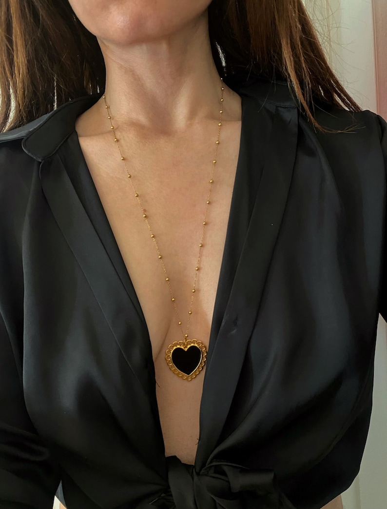 Pu&ograve; includere: Una collana con catena dorata con un ciondolo a forma di cuore nero. Il ciondolo ha un design dettagliato e decorato attorno al bordo.