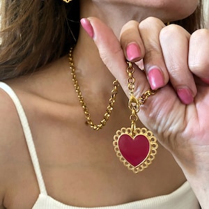 May include: A gold chain necklace with a large red heart pendant. The pendant has a delicate, scalloped edge. The necklace is paired with matching earrings featuring smaller red heart charms.
