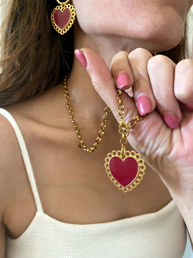 May include: A gold chain necklace with a large red heart pendant. The pendant has a delicate, scalloped edge. The necklace is paired with matching earrings featuring smaller red heart charms.