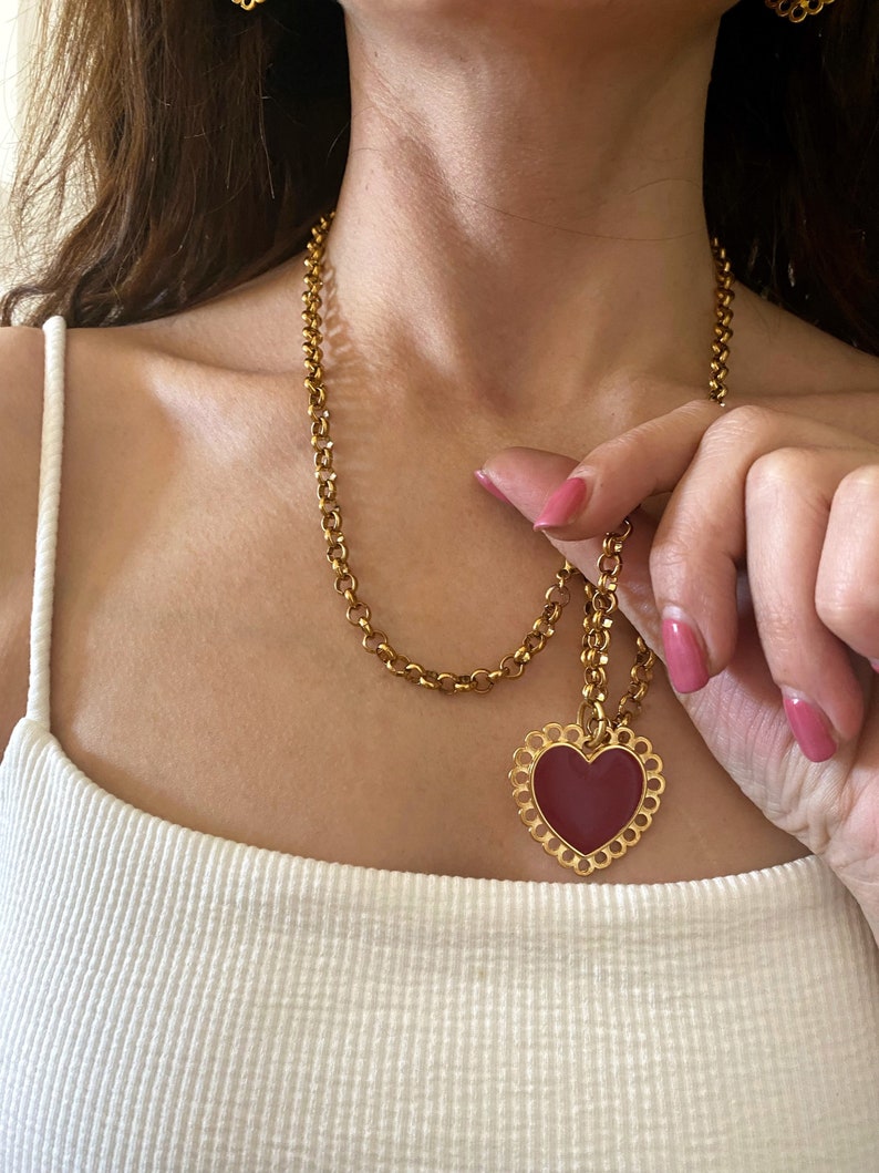 May include: A gold chain necklace with a large, red heart pendant. The heart has a delicate, ornate border.
