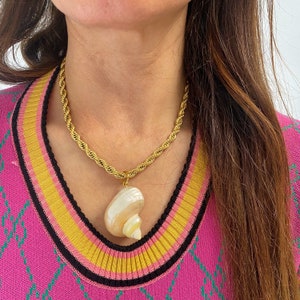 May include: A gold rope chain necklace with a large, white seashell pendant. The necklace is worn by a person with long brown hair and a pink and yellow striped sweater.