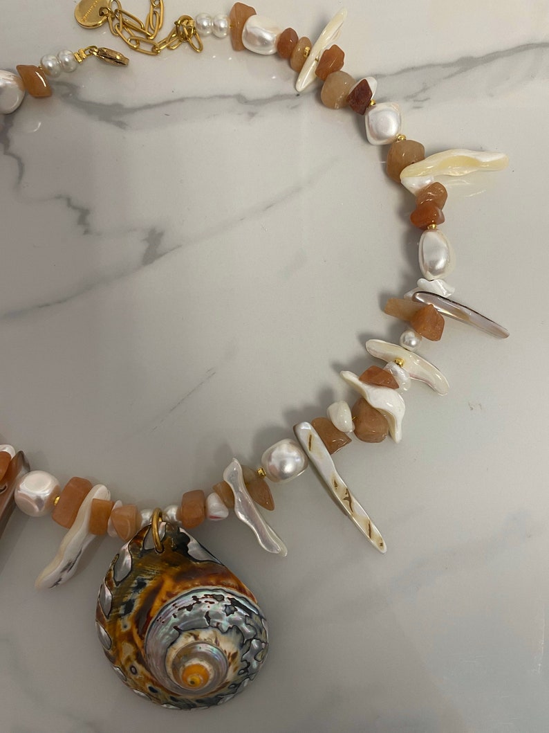 May include: A gold chain necklace with a large seashell pendant. The necklace is made with white pearls, brown and orange stones, and white and brown shell pieces.
