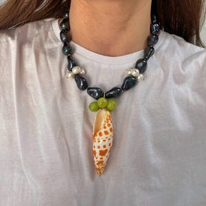 May include: A black and iridescent pearl necklace with a shell pendant. The shell is orange and white with brown spots and is surrounded by green beads.