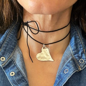 Può includere: Una collana con pendente a cuore argentato su un choker in cordoncino nero. Il pendente a forma di cuore ha una texture martellata e pende da un delicato cordoncino nero che si annoda dietro il collo. La collana è indossata sopra una giacca di jeans.