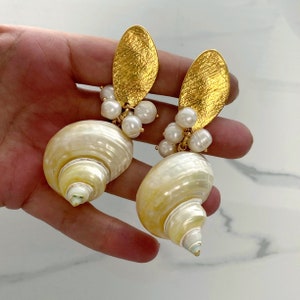 Könnte beinhalten: Ein Paar goldene und weiße Muschelohrringe mit Perlenakzenten. Die Ohrringe haben eine große, weiße Muschel mit einem goldenen, blattartigen Anhänger und einem Perlenschmuck.