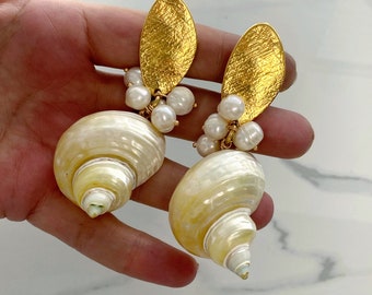 Könnte beinhalten: Ein Paar goldene und weiße Muschelohrringe mit Perlenakzenten. Die Ohrringe haben eine große, weiße Muschel mit einem goldenen, blattartigen Anhänger und einem Perlenschmuck.