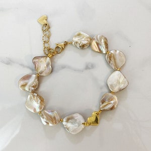 Puede incluir: Un brazalete dorado con un dije de corazón y una cadena de perlas de color blanco roto de forma irregular.