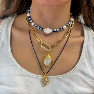 K&ouml;nnte beinhalten: Ein mehrlagiges Halsketten-Set mit einer goldenen Kette mit einem gro&szlig;en Barockperlen-Anh&auml;nger, einer goldenen Kette mit einem rechteckigen Anh&auml;nger und einem blauen b&ouml;sen Blick-Perlen-Choker.