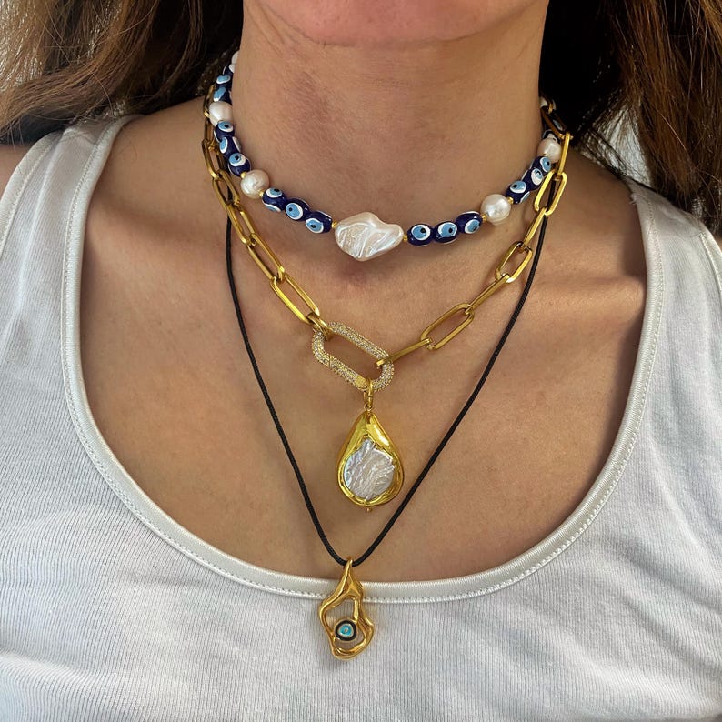 K&ouml;nnte beinhalten: Ein mehrlagiges Halsketten-Set mit einer goldenen Kette mit einem gro&szlig;en Barockperlen-Anh&auml;nger, einer goldenen Kette mit einem rechteckigen Anh&auml;nger und einem blauen b&ouml;sen Blick-Perlen-Choker.