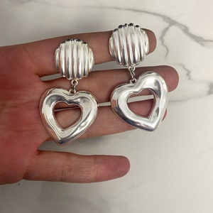 May include: Silver heart-shaped earrings with a textured, ridged design. The earrings have a cutout heart shape in the center.