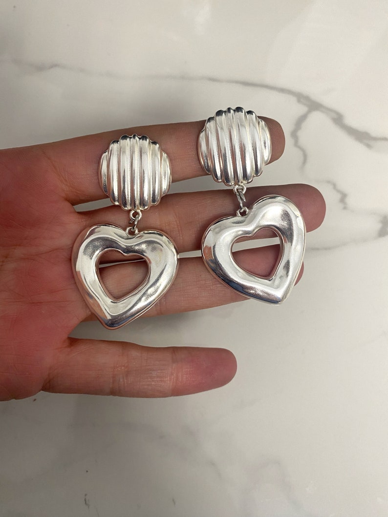 May include: Silver heart-shaped earrings with a textured, ridged design. The earrings have a cutout heart shape in the center.