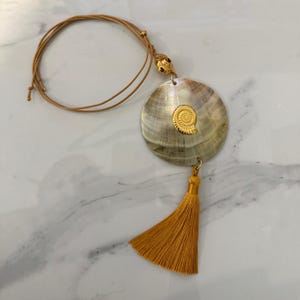 Puede incluir: Un collar con un colgante redondo de nácar iridiscente adornado con un pequeño dije de fósil de ammonite en tono dorado. El colgante cuelga de un cordón fino de color tostado y está acentuado con una borla de tela amarillo mostaza.