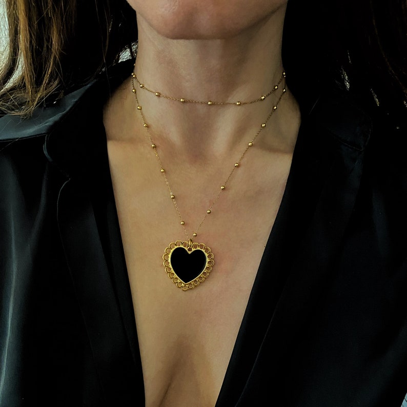 Pu&ograve; includere: Una collana con catena dorata con un ciondolo a forma di cuore nero. Il ciondolo ha un design delicato e decorato.