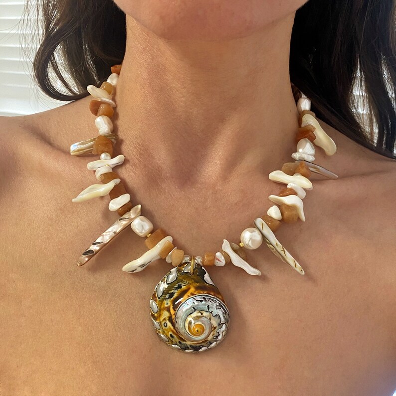 May include: A brown and white shell necklace with a large, spiral-shaped shell pendant. The necklace is made of various sizes and shapes of shells, including some that are pointed.