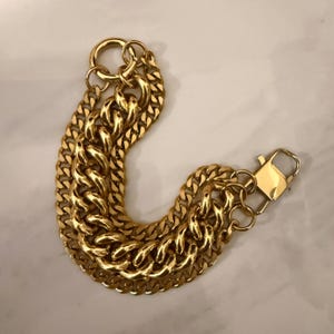 Könnte beinhalten: Ein goldfarbenes Kettenarmband mit zwei dicken, ineinandergreifenden Panzerkettensträngen. Das Armband verfügt über einen großen kreisförmigen Ring an einem Ende und einen rechteckigen Karabinerverschluss am anderen Ende mit einer polierten, reflektierenden Oberfläche.